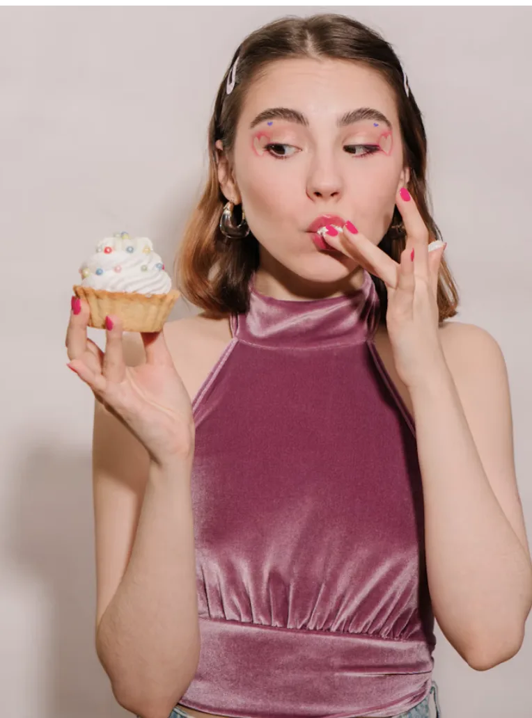 Woman in a mauve velvet top licking frosting from her finger while holding a cupcake